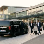 Minibus premium noir stationné devant un terminal d'aéroport avec un groupe de voyageurs